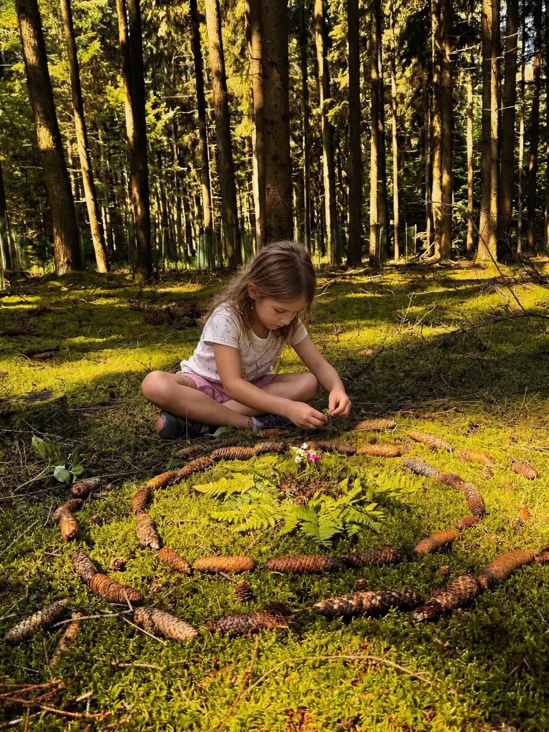 Ein Mädchen legt im Wald eine Spirale aus Zapfen und Stöcken auf das Moos
